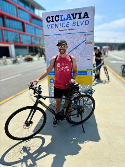 Man smiling with a bicycle in front of a CicLAvia Venice Blvd event sign on a sunny day, wearing a red tank top, black shorts, sunglasses, and a cap.